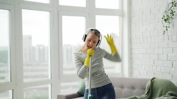 快乐的年轻金发女郎在现代房子里做家务和娱乐，她用拖把洗地板，唱歌和跳舞通过耳机听收音机。