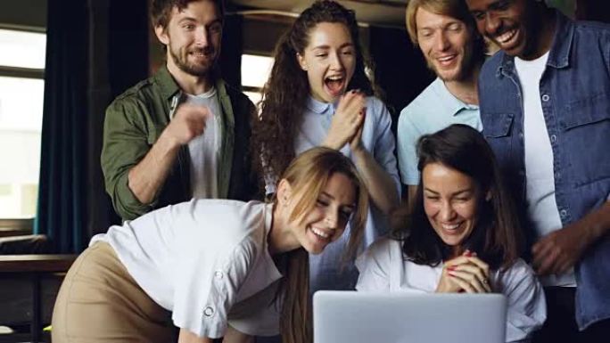 漂亮的黑发女人正在使用计算机并大笑，她的团队来祝贺她取得了良好的成绩，人们在鼓掌和交谈。
