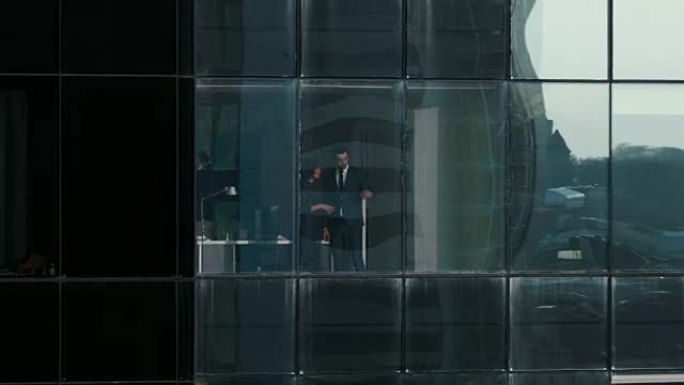 摩天大楼外的航拍镜头: 商人和女商人站在办公室窗户旁谈论生意。金融商务区和在大城市工作的商人的飞行镜