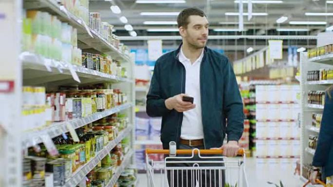 在超市: 英俊的男人使用智能手机，看着罐头食品的营养价值。他站在罐头食品区的购物车。
