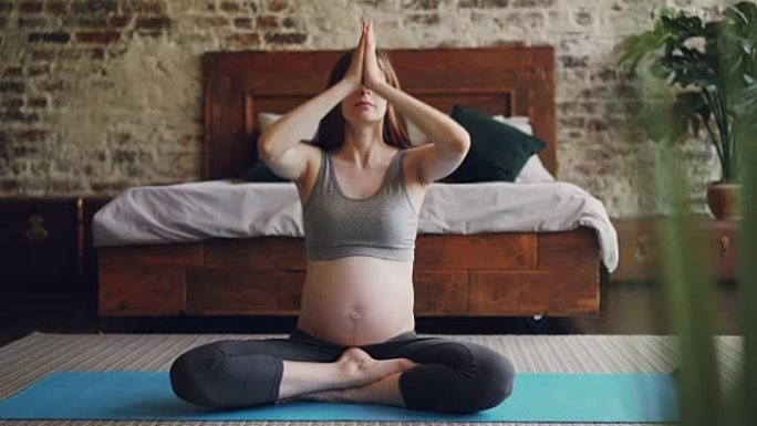 好看的怀孕女孩正在家里以莲花姿势冥想，坐在瑜伽垫上举起手臂，然后将手掌放在膝盖上放松。幸福和怀孕概念