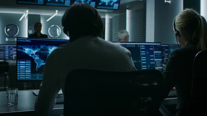 在个人计算机上工作的网络安全调度员的后视图，显示了充满特殊情报人员的系统控制室中的交通数据流。