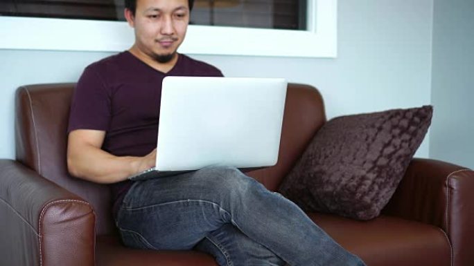 4k镜头亚洲男子休闲西装坐在沙发上思考与科技笔记本电脑一起工作的场景早上在卧室，生活方式和休闲概念