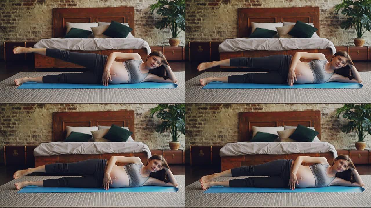 健康的孕妇抬起腿躺在卧室地板上的瑜伽垫上做体育锻炼。健康的生活方式，期待母亲和室内设计的概念。