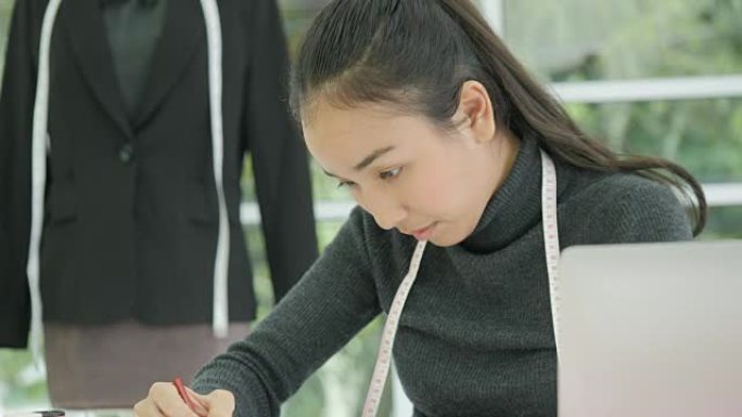 美丽的亚洲女性时装设计师坐在办公桌前看着现代工作室笔记本电脑上的草图，做笔记