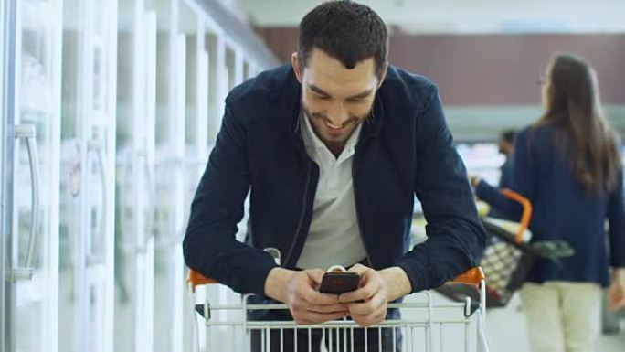 在超市: 英俊的男人用智能手机微笑，同时靠在购物卡上。与其他人一起在商店的冰冻部分购物的顾客快乐。