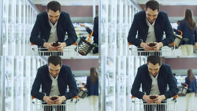 在超市: 英俊的男人用智能手机微笑，同时靠在购物卡上。与其他人一起在商店的冰冻部分购物的顾客快乐。