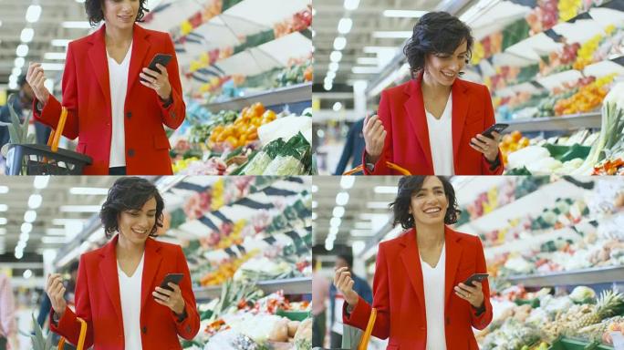 在超市: 美丽的女人使用智能手机，走过商店的新鲜农产品区。女人沉浸在网上冲浪在她的手机背景五颜六色的