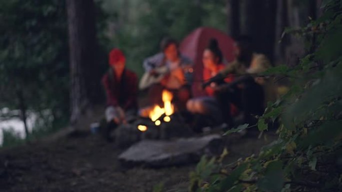模糊的镜头是游客朋友坐在森林的篝火旁，弹吉他并在火炉旁唱歌。关注前景中的树枝。