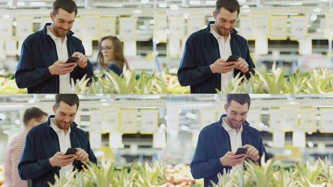在超市: 英俊的男人站在新鲜农产品区附近选择有机水果时，使用智能手机，浏览互联网并迷人地微笑。