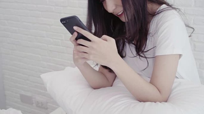 慢动作-美丽的亚洲女人在卧室躺在床上时使用智能手机。快乐的女性在家中使用手机发短信，阅读，短信和在线