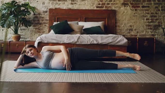 漂亮的怀孕女孩正在做体育锻炼，抬高腿躺在卧室地板上的瑜伽垫上，背景是阁楼风格的房间和木制家具。