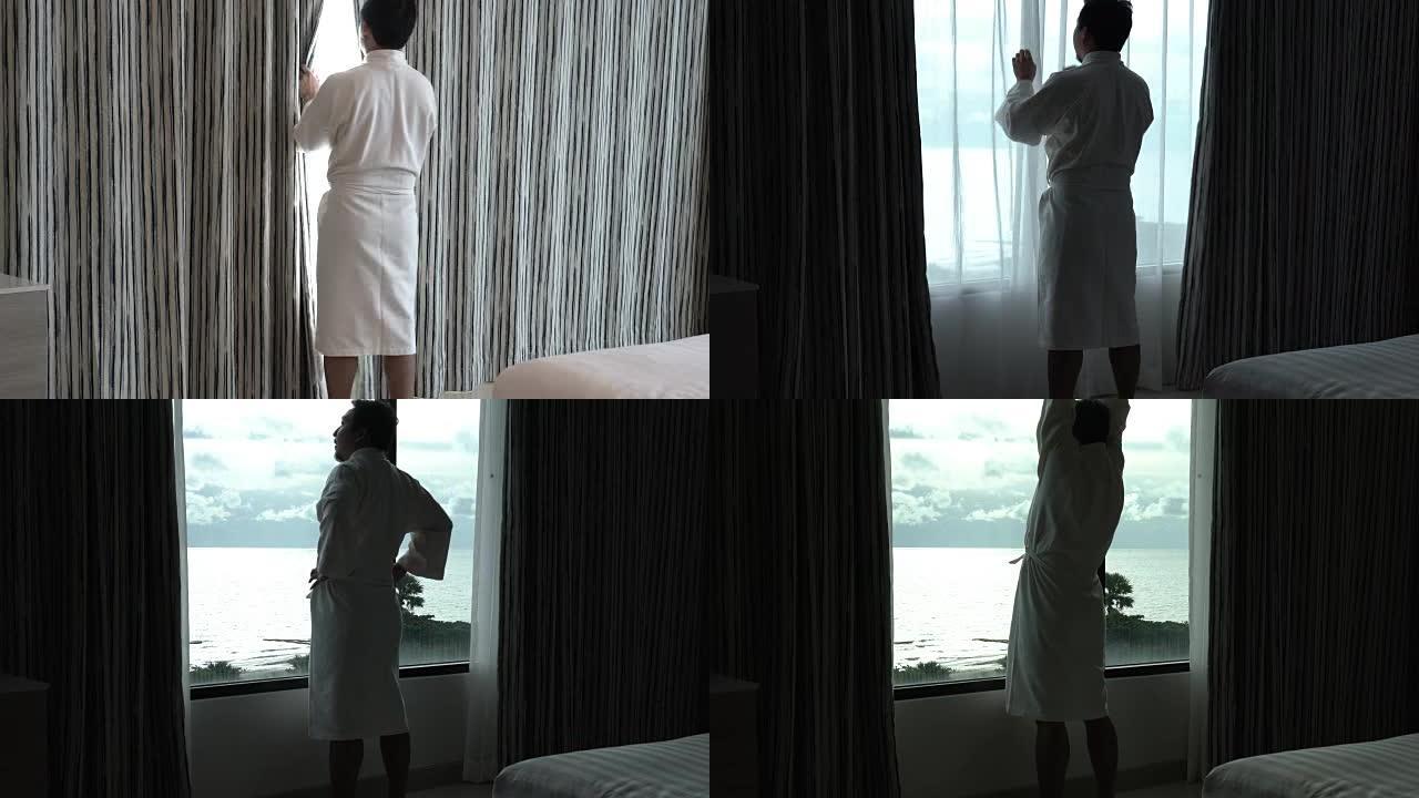 4k镜头亚洲男子穿着浴袍散步的场景，早上在豪华酒店卧室醒来时打开窗户上的窗帘，生活方式和休闲概念