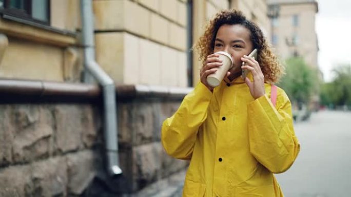 漂亮的非裔美国妇女正在喝咖啡，在手机上聊天，穿着明亮的夹克在春天的城市散步。现代技术和千禧一代的概念