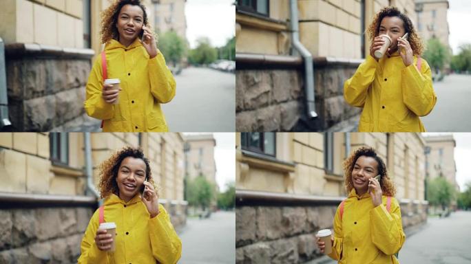漂亮的非裔美国妇女正在喝咖啡，在手机上聊天，穿着明亮的夹克在春天的城市散步。现代技术和千禧一代的概念