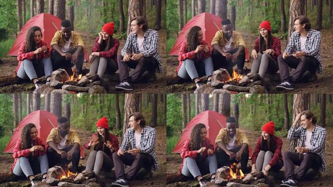 快乐的游客男人和女人坐在篝火旁分享故事，在燃烧的火焰旁笑着变得温暖。对话、人和有趣的概念。