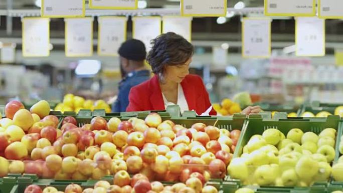 在超市: 美女在农贸市场的新鲜农产品区选择有机水果。她拿起水果，把它们放进购物篮。慢动作。