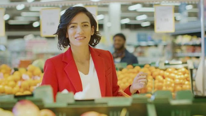 在超市: 美丽微笑的女人在新鲜农产品过道中选择产品并将其放入购物篮的肖像。在背景五颜六色的水果和有机