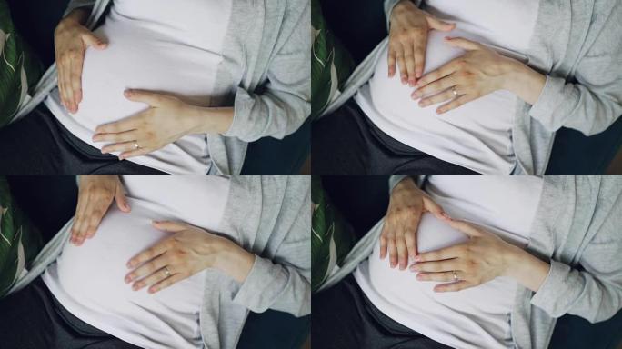 怀孕的腹部和温柔的女性双手的特写镜头用温柔和关怀抚摸和抚摸婴儿bump，然后用手指使心脏象征着对孩子