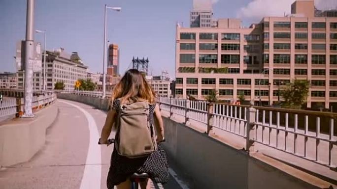 后视图女通勤骑自行车与背包和金色头发在风中吹在一个阳光明媚的日子慢动作
