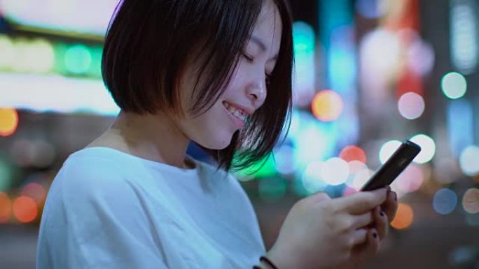 带有穿孔和穿着休闲服的迷人日本女孩的肖像使用智能手机。在后台，大城市的广告广告牌灯光在夜晚发光。