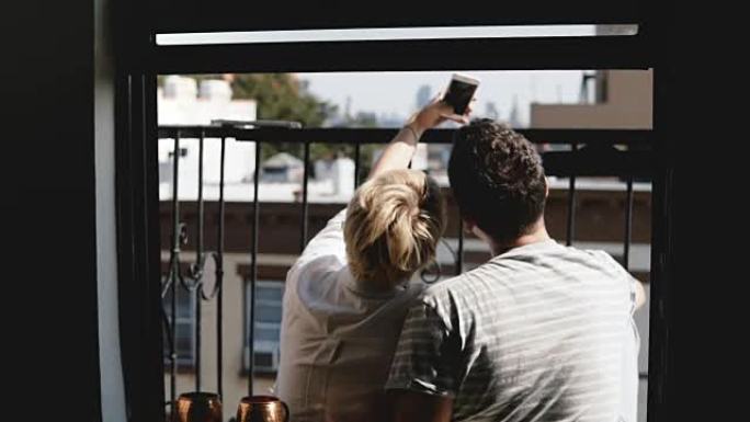 后视图快乐的年轻夫妇坐在纽约一个阳光明媚的夏季公寓阳台上，拍摄智能手机自拍