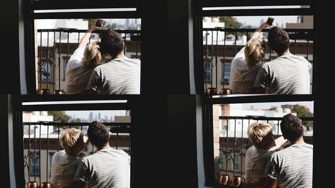 后视图快乐的年轻夫妇坐在纽约一个阳光明媚的夏季公寓阳台上，拍摄智能手机自拍