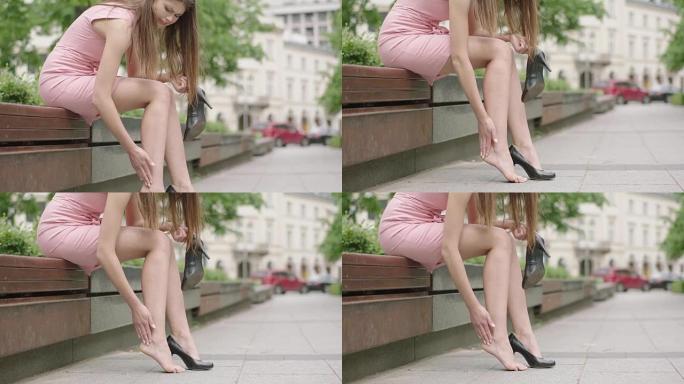 穿着粉色连衣裙的疲惫年轻女子坐在长凳上按摩脚