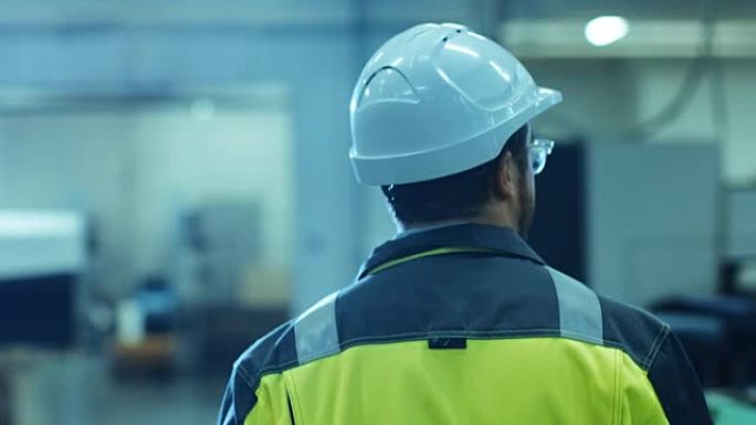 在背面拍摄的穿着防护服的工业工程师戴上安全帽，走进现代制造设施，背景是自动化机械。