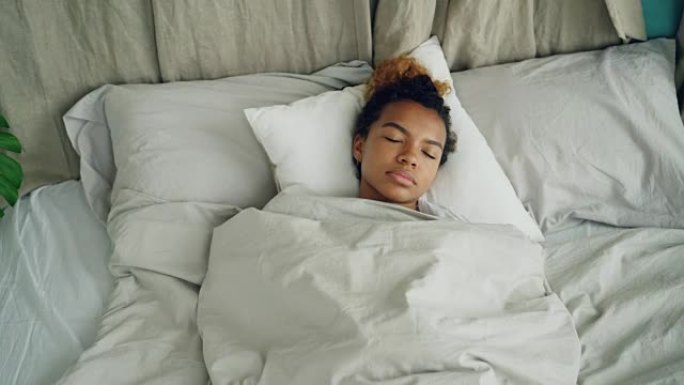 年轻的非洲裔美国妇女睡在舒适的床上，在温暖的毯子下，在漂亮的亚麻布上休息。舒适、休息的人和卧室概念。