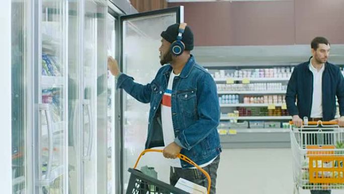 在超市: 戴着耳机的时尚非洲裔美国人从冰箱中选择冷冻商品中的产品，并将其放入购物篮。慢动作。