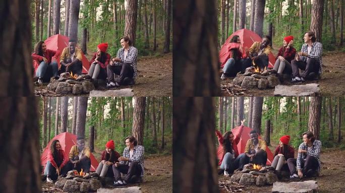 快乐的女孩和男孩的朋友在营地里取暖，在火炉周围聊天，打手势和大笑。对话、自然和人的概念。
