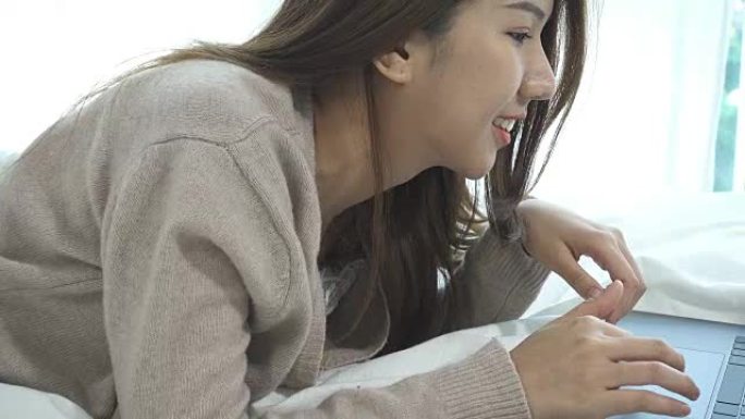美丽的年轻微笑的亚洲女人，坐在家里卧室的床上，在笔记本电脑上工作。亚洲女商人在家里放松。在家享受时光