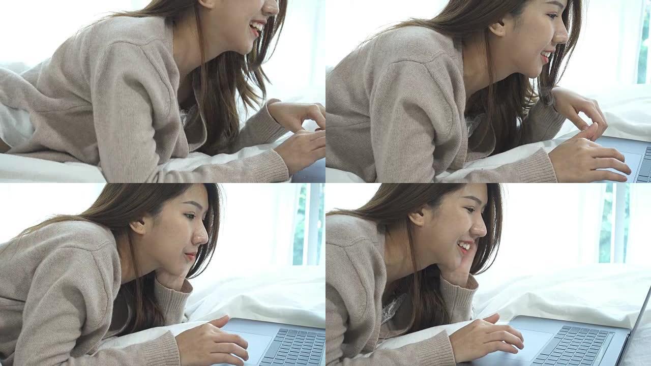 美丽的年轻微笑的亚洲女人，坐在家里卧室的床上，在笔记本电脑上工作。亚洲女商人在家里放松。在家享受时光