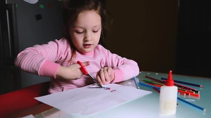 可爱的小学龄前欧洲女孩孩子穿着粉色毛衣把她的画从纸上剪出来，和某人说话