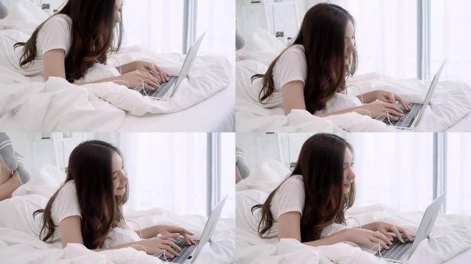 美丽的亚洲女人躺在卧室的床上玩电脑。快乐的女性在家中戴着耳机听音乐并在笔记本电脑中搜索歌曲。生活方式