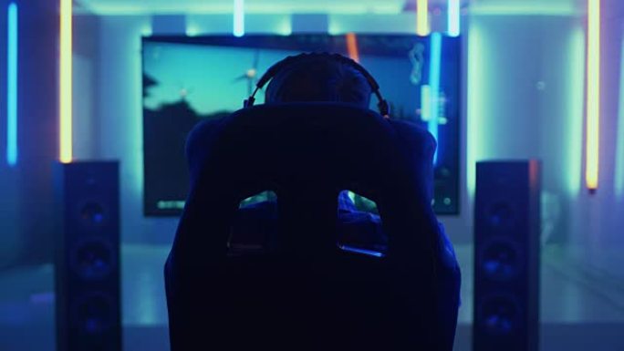 后视图镜头的职业玩家坐在赛车座椅模拟器驾驶舱与方向盘和玩赛车在线视频游戏。大电视和背景为霓虹灯的房间