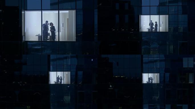 摩天大楼外的航拍画面: 商人和女商人站在办公室的窗户里聊天。晚上检索商业区的飞行镜头和在大城市工作的