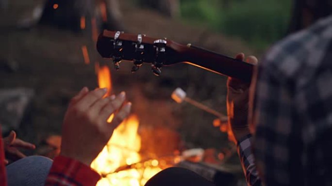 男性手弹吉他和女性手在背景中燃烧篝火跳舞的特写镜头。露营、音乐和人的概念。