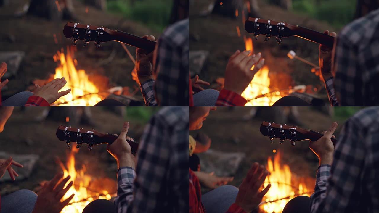 男性手弹吉他和女性手在背景中燃烧篝火跳舞的特写镜头。露营、音乐和人的概念。