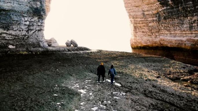 无人机跟随快乐的旅游夫妇沿着诺曼底史诗巨拱下令人难以置信的雪花石膏低潮海滩散步。