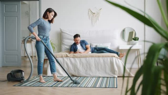 妻子在卧室吸尘地毯，而丈夫在床上使用智能手机