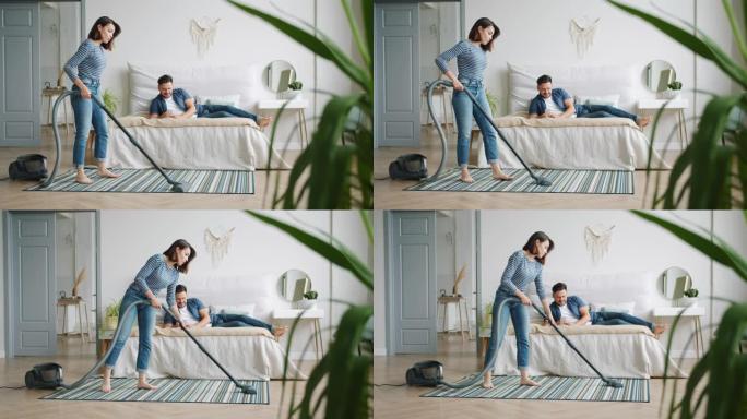 妻子在卧室吸尘地毯，而丈夫在床上使用智能手机