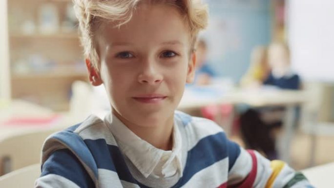 一个可爱的小男孩的肖像，波浪形的金发坐在他的课桌前，开心地微笑着。聪明的小男孩，带着迷人的微笑坐在教