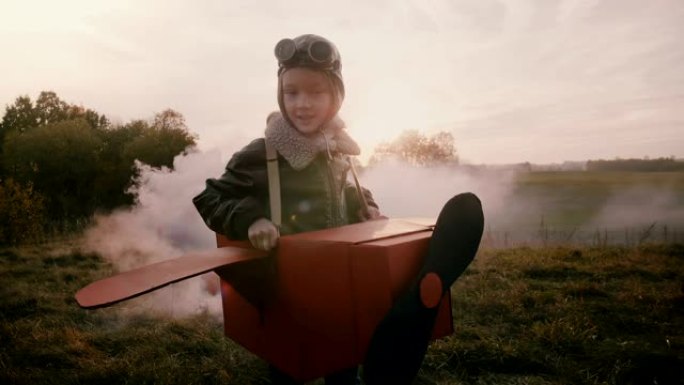 小飞行员女孩在有趣的纸板飞机上奔跑，背后是彩色烟雾，在惊人的秋季日落场慢动作。