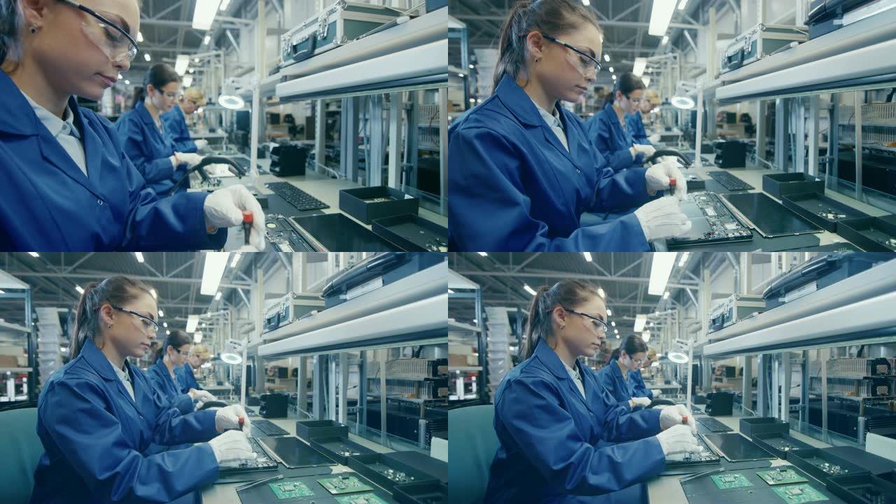 穿着蓝色工作服和防护眼镜的女电子工厂工人正在用螺丝刀组装笔记本电脑的主板。拥有多名员工的高科技工厂设