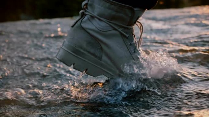 日落慢动作时，穿着灰色防水沙漠鞋的男性腿的特写镜头直接走进水流。