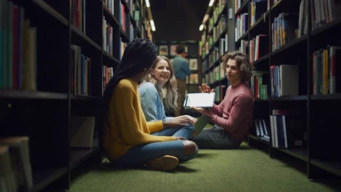大学图书馆: 各种各样的学生盘腿坐在地板上，交谈，使用数字平板电脑，共享屏幕，玩得开心，讨论论文，一