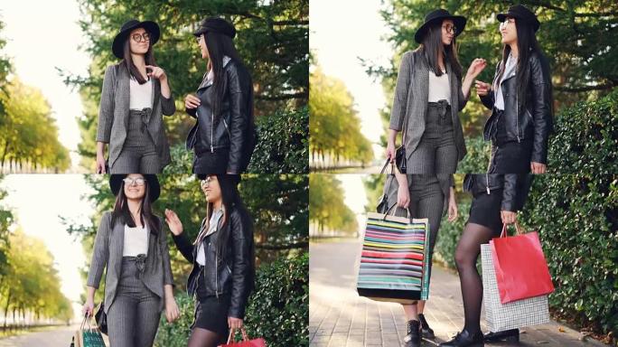 快乐的女孩站在户外人行道上聊天，笑着拿着明亮的购物袋在购物中心讨论一天。女人穿着时髦的衣服和眼镜。