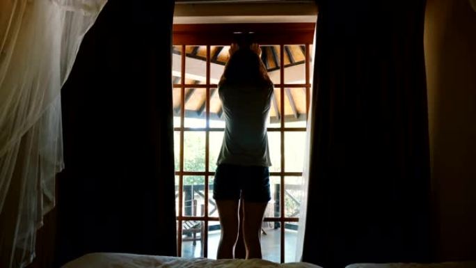 后视图美丽的年轻幸福的女人打开酒店小屋窗帘和窗户门，走到令人惊叹的阳光明媚的露台。